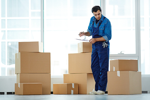 Service worker in uniform looking at one of boxes in stack while reading list of delivered things