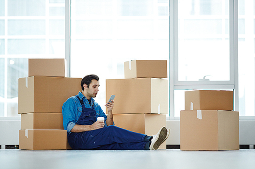 Restful worker in uniform sitting on the floor while leaning against stack of boxes and messaging in smartphone