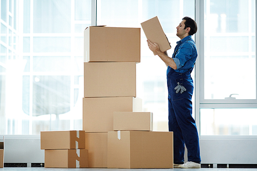 Young relocation agency staff in workwear going to put box on top of stack while doing his work