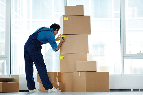 Delivery man in uniform sticking notepapers with numbers on packed boxes before loading
