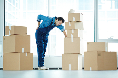 Young relocation service staff touching his back and leaning by stack of packed boxes with supplies during work