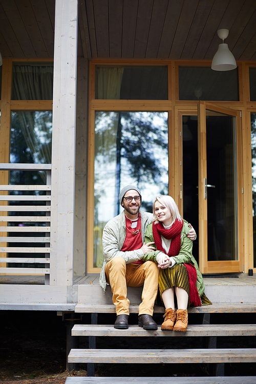 Portrait of young beautiful stylish Caucasian couple sitting on front stoop of new house and smiling at camera happily