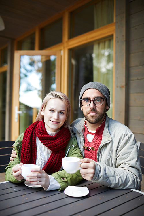 Portrait of young attractive Caucasian couple sitting at wooden table on terrace, holding cups of hot tea and 