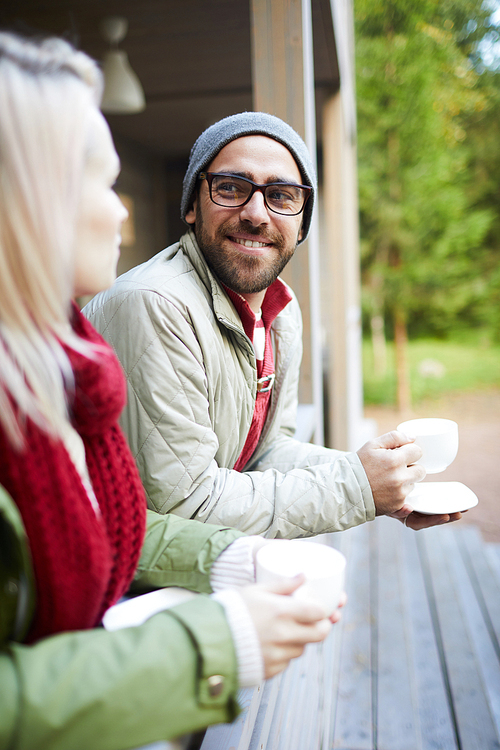 Attractive bearded Caucasian man talking to beloved and smiling cheerfully while standing on porch with cup of fresh tea on autumn afternoon