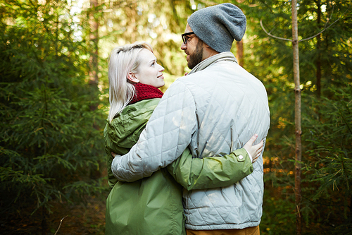 Rear view of young loving Caucasian couple embracing each other and looking at each other with admiration while walking in woods of pine