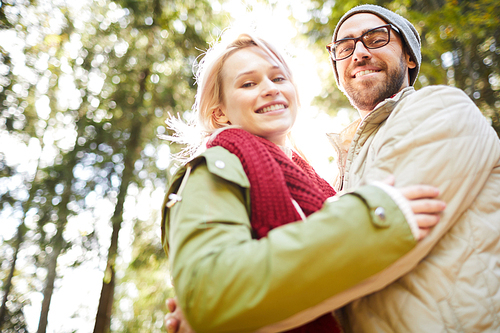 Low angle view of young Caucasian couple embracing each other and smiling at camera cheerfully while walking in park on sunny autumn day