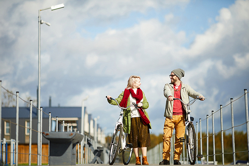 Young stylish Caucasian couple chatting and smiling happily while walking along the pier with bicycles on warm autumn afternoon, low angle view