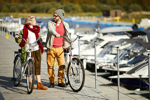 Young stylish Caucasian couple walking down the pier with bikes, chatting and enjoying gorgeous sunny autumn afternoon