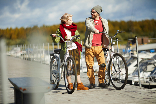 Handsome bearded man talking to young pretty woman while walking down the pier with bicycles on warm bright autumn afternoon