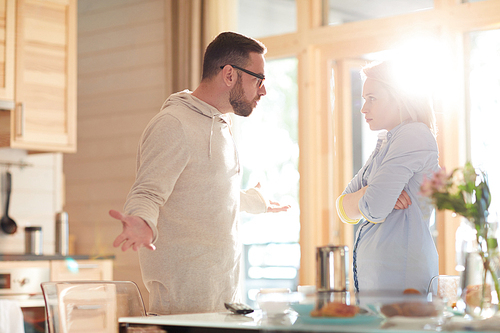 Young married Caucasian couple arguing about household duties while standing in home kitchen illuminated with bright morning sun