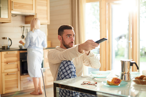 Young bearded man sitting at kitchen table and switching channels on TV with remote control while his wife making breakfast