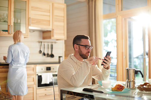 Bearded Caucasian man in glasses sitting at kitchen table and texting on smartphone while his wife washing dishes after breakfast