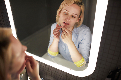 Young attractive blond Caucasian woman popping pimple on her chin while looking at herself in bathroom mirror