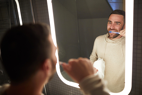 Reflection of young bearded Caucasian man brushing his teeth with toothbrush and looking at himself in bathroom mirror