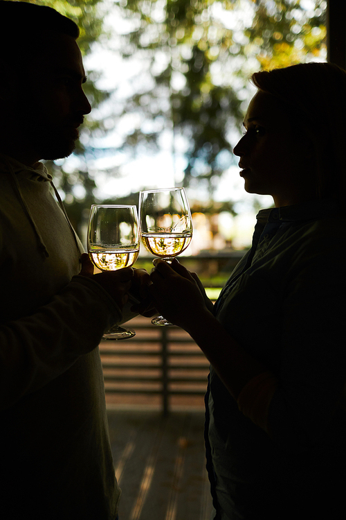 Silhouette of unrecognizable amorous couple clinking glasses of white wine and looking at each other with love while standing on terrace