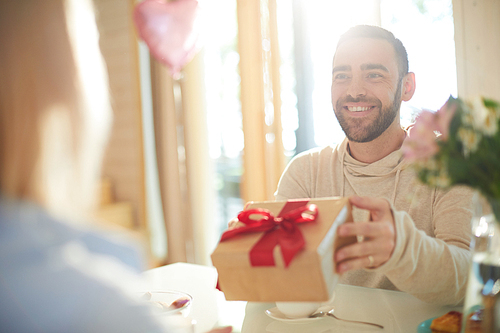 Young handsome bearded Caucasian man presenting gift to his girlfriend and smiling cheerfully while celebrating Valentines Day