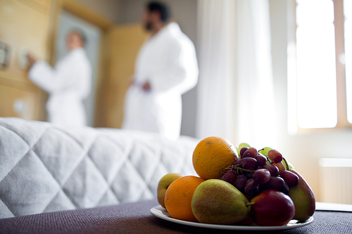 Pile of fresh fruits on plate on small table in room of new house with couple in bathrobes on background