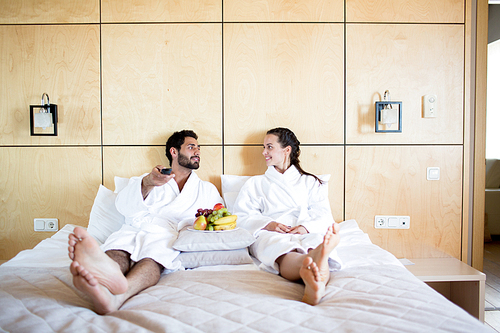 Young restful couple in bathrobes relaxing on bed, having fruits and choosing what to watch on tv
