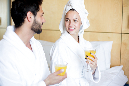 Young couple with glasses of orange or peach juice having talk in bedroom or hotel room on vacation