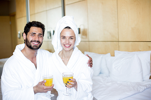 Happy young affectionate couple in bathrobes sitting in bedroom and having juice after bath