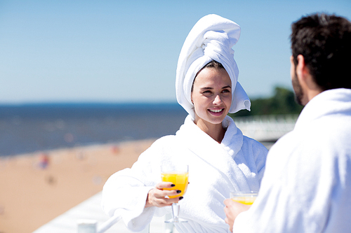 Young woman in white bathrobe and towel having juice and talking to her husband during honeymoon vacation at summer resort