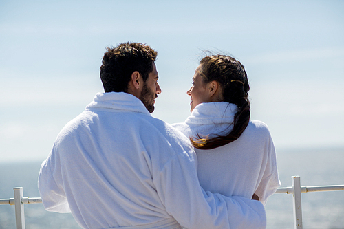 Back view of man in bathrobe embracing his wife during talk by waterside at summer resort