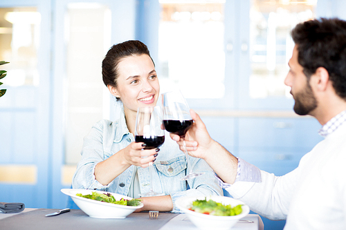 Young amorous couple clinking with red wine while celebrating family occasion in restaurant