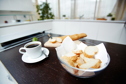 Slices of fresh wheat baguette with sesam in bowl and cup of black coffee on table in the kitchen