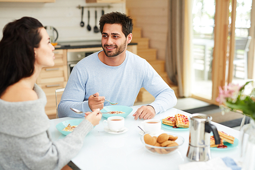 Cheerful positive young multi-ethnic couple in homewear sitting at table and eating healthy breakfast while chatting at home