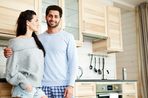 Cheerful handsome young bearded man in pajamas smiling at camera and hugging his girlfriend while they standing in cozy kitchen with wooden set