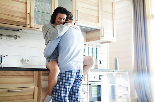 Content sensual lovers in casual clothing hugging in domestic kitchen: young woman sitting on kitchen set and keeping eyes closed while cuddling with tenderness beloved man