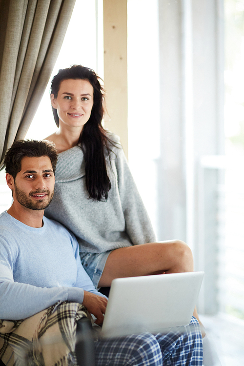 Smiling beautiful young couple in comfortable homewear sitting on one armchair and  while resting with laptop at home