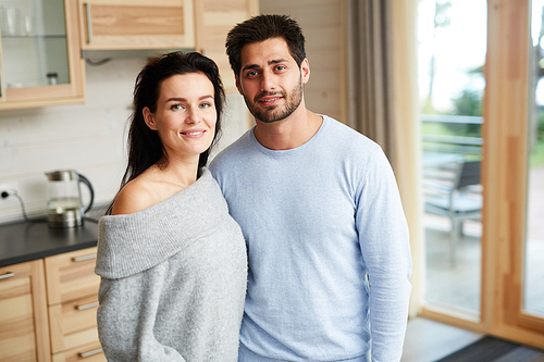 Positive attractive young couple in homewear standing in rustic kitchen and smiling at camera