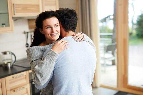 Happy smiling beautiful brunette woman hugging boyfriend  and looking at view while they standing in cozy kitchen at home