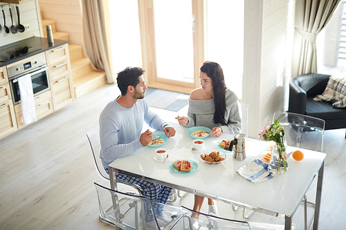 Serious young couple sitting at dining table full of desserts and eating breakfast while talking in kitchen