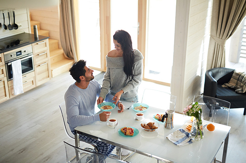 Smiling pretty young wife in gray sweater taking care of husband and putting plate of muesli on table while talking to him at home