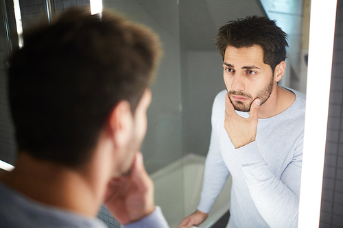 Serious handsome brunette young man touching beard and looking into mirror while thinking about shaving in bathroom