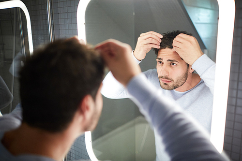 Serious frowning young man viewing his hair and finding receding hairline while looking into mirror and touching hair, he suffering from hair loss