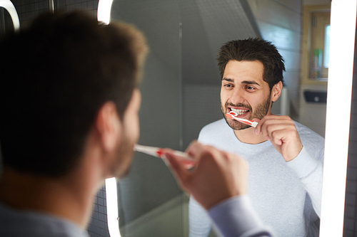 Cheerful handsome young bearded man looking into illuminated mirror in bathroom and cleaning teeth with toothbrush in morning