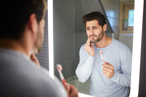 Sad frowning handsome brunette young man with beard touching cheek and looking into mirror in bathroom while getting bad tooth during cleaning teeth