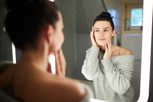 Smiling attractive young lady in sweater looking into mirror and touching face while applying cream on cleaned face in bathroom