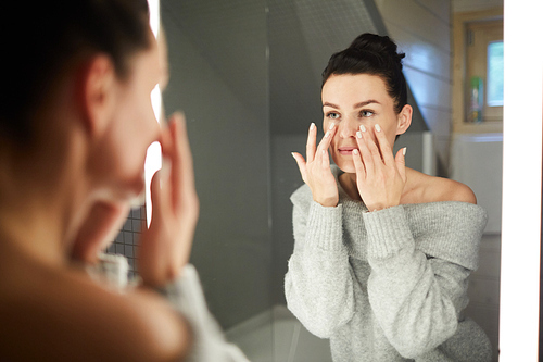 Content attractive brunette young woman in gray sweater looking into mirror and massaging face while applying cream in bathroom