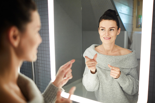 Cheerful confident cool young woman wearing nude off shoulder sweater pointing finger guns and looking into mirror in modern bathroom