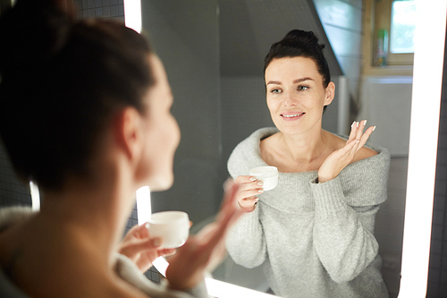 Happy attractive young woman with fresh skin looking into mirror and applying cream on face in bathroom, she taking care of face skin