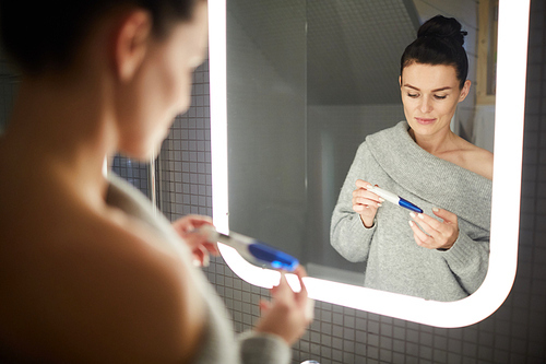 Serious calm young woman with hair bun standing against illuminated mirror in bathroom and checking pregnancy test