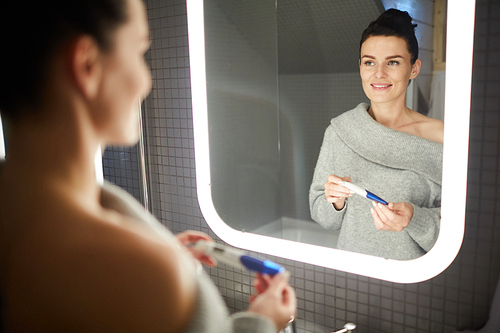 Happy excited young woman in nude off shoulder sweater looking into mirror and smiling while receiving positive pregnancy test in bathroom