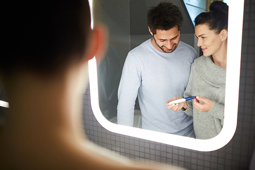 Content attractive young wife showing pregnancy test to husband while sharing good news in bathroom