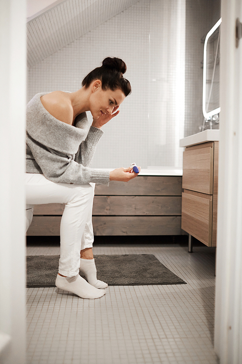 Upset shocked attractive young woman with hair bun sitting on toilet bowl in bathroom and looking at pregnancy test, she not believing her eyes