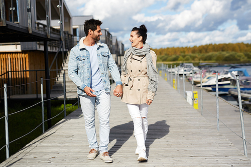 Cheerful excited young couple in stylish clothes holding hands and walking together on pier while chatting with each other