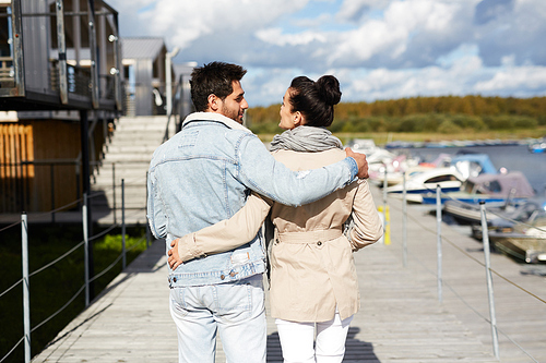 Rear view of young couple in casual clothing embracing each other and enjoying stroll on warm autumn day while walking on pier